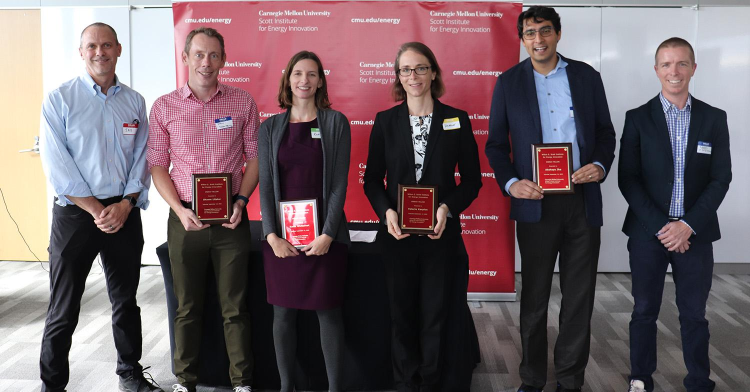 Six people stand and pose for the photograph. The middle four hold awards.