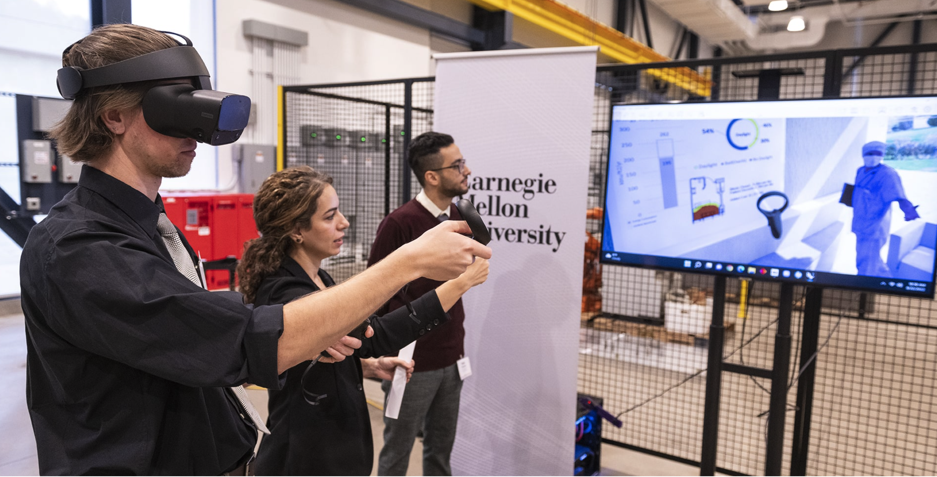 Students try out a virtual reality experience. One wears a headset and holds a controller while two other people watch a television.
