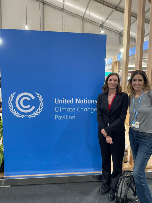Katie Whitefoot and Paulina Jaramillo stand next to a tall banner which reads "United Nations Climate Change Pavilion."